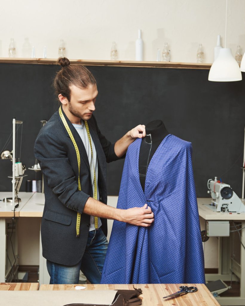 Young shaved good-looking caucasian male fashion designer in stylish outfit working on new blue dress for spring collection in his workshop. Artist creating beautiful clothes in his workshop.